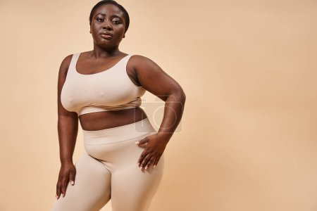 plus size afrikanisch-amerikanische Frau in Unterwäsche schaut in die Kamera auf beigem Hintergrund, Körper positiv