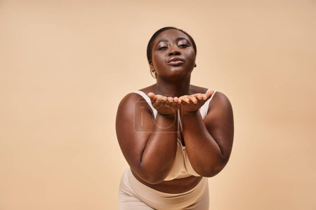 junge plus size afrikanisch-amerikanische Frau in beiger Unterwäsche bläst Luftkuss vor passender Kulisse