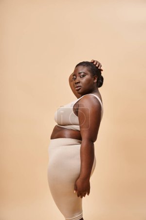 young plus size african american woman in neutral color underwear posing against matching backdrop