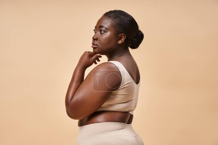 pensive plus size african american woman in beige underwear posing against matching backdrop