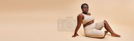 plus la taille afro-américaine jeune femme en sous-vêtements beige posant sur fond assorti, bannière