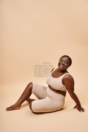 happy plus size african american young woman in beige underwear posing against matching backdrop