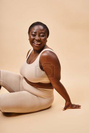 fröhliche afrikanisch-amerikanische junge Frau in beiger Unterwäsche posiert vor passender Kulisse
