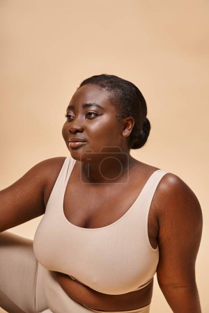 verträumtes Plus Size afrikanische junge Frau in beiger Unterwäsche posiert vor passendem Hintergrund