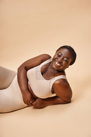 happy plus size african american young woman in beige lingerie reclining against matching backdrop