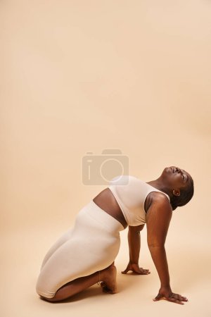 modelo afroamericano de talla grande en lencería beige riéndose y reclinándose contra fondo a juego