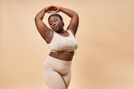 plus size african american woman in underwear posing with raised hands against beige backdrop