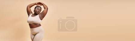 plus size african american woman in underwear posing with raised hands on beige backdrop, banner