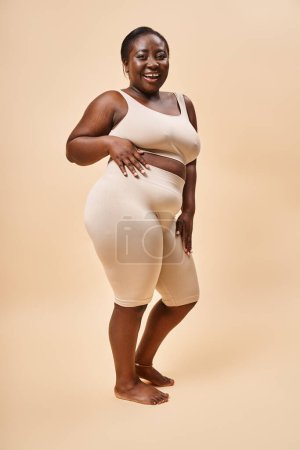 Selbstbewusste Plus-Size-Frau in beiger Unterwäsche posiert mit verschränkten Armen, positivem Körper und Selbstwertgefühl