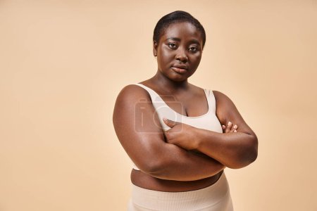 Plus Size Frau in beiger Unterwäsche posiert mit verschränkten Armen, positivem Körper und Selbstwertgefühl