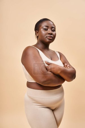 glückliche Plus Size Frau in beiger Unterwäsche posiert mit verschränkten Armen, positivem Körper und Selbstwertgefühl