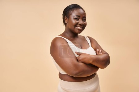 glückliche Plus-Size-Frau in beiger Unterwäsche posiert im Studio, Körper positiv und Selbstwertgefühl