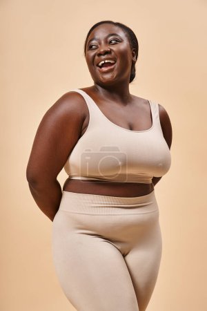 glückliche Plus-Size-Frau in beiger Unterwäsche lacht im Studio, Körper positiv und Selbstwertgefühl