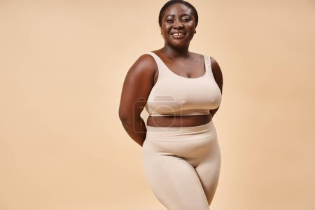 fröhliche Plus-Size-Frau in beiger Unterwäsche posiert im Studio, Körper positiv und Selbstwertgefühl