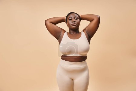 calm plus size woman in beige underwear posing with hands near head, body positive and serenity