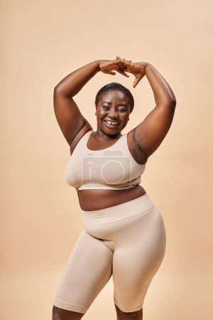 glückliche Plus Size Frau in beiger Unterwäsche posiert mit erhobenen Händen, positivem Körper und Selbstwertgefühl