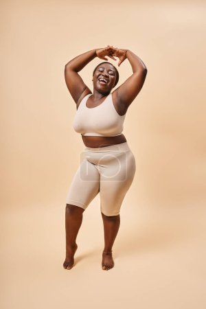 glückliche Plus Size Frau in beiger Unterwäsche posiert mit erhobenen Händen, positivem Körper und Selbstwertgefühl