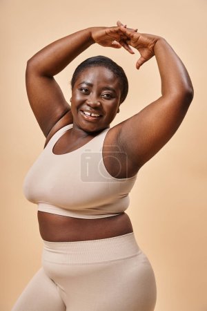 lächelnde Plus Size Frau in beiger Unterwäsche posiert mit erhobenen Händen, positivem Körper und Selbstwertgefühl