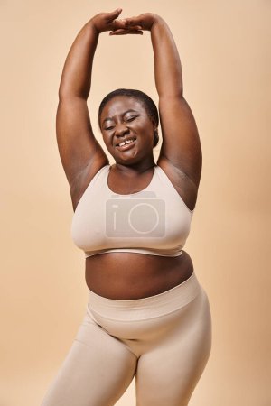 freudige Plus-Size-Frau in beiger Unterwäsche posiert mit erhobenen Händen, positivem Körper und Selbstwertgefühl
