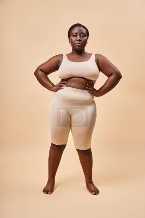Plus Size Frau in beiger Unterwäsche posiert mit den Händen auf Hüften, Body Positive und Female Empowerment
