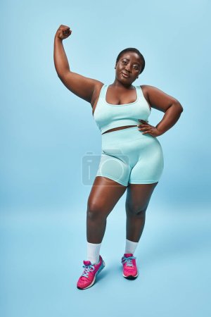 Energetic plus size woman in active wear flexing her muscles on blue background, hand on hip
