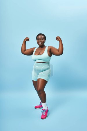 starke Plus Size Frau in aktiver Kleidung lässt ihre Muskeln spielen und schaut in die Kamera auf blauem Hintergrund