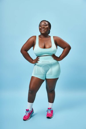 happy plus size african american woman in blue sportswear with hands on hips on matching backdrop