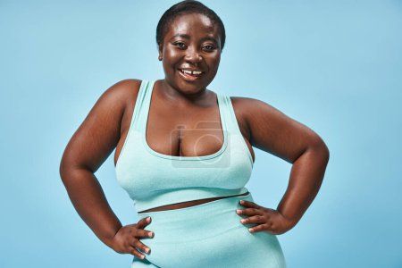 heureux plus la taille femme afro-américaine en bleu vêtements de sport avec les mains sur les hanches sur fond assorti