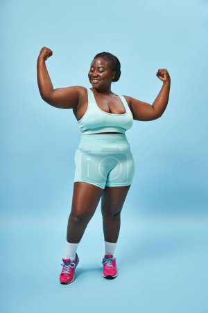 fröhliche Plus-Size-Frau in aktiver Kleidung, die ihre Muskeln spielen lässt und auf blauem Hintergrund lächelt