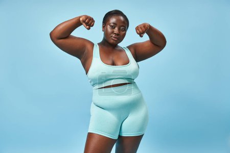 mujer seria de talla grande en ropa activa flexionando sus músculos y mirando a la cámara sobre fondo azul