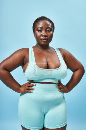 fuerte más tamaño mujer afroamericana en ropa deportiva azul con las manos en las caderas en el telón de fondo a juego