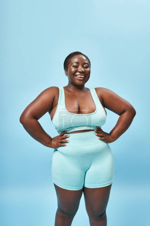 mujer afroamericana feliz en ropa deportiva azul con las manos en las caderas en el telón de fondo a juego