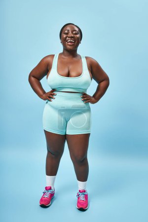 jolly plus size african american woman in blue sportswear with hands on hips on matching backdrop