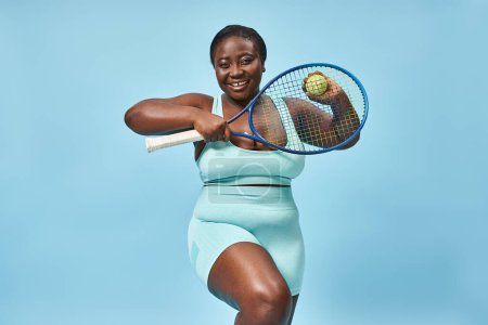 happy plus size african american woman ready to play tennis on blue background, body positive