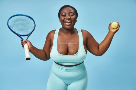 smiling plus size african american woman holding ball and tennis racket on blue background, sport