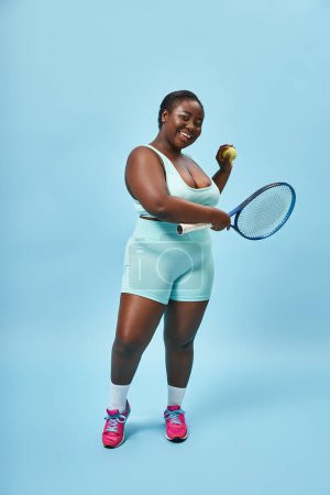 lächelnd plus size afrikanisch amerikanisch frau bereit, tennis zu spielen auf blauem hintergrund, körper positiv