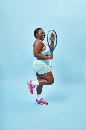 alegre más tamaño de piel oscura mujer sosteniendo raqueta de tenis sobre fondo azul, cuerpo positivo y deporte