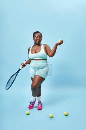 Photo for Happy plus size dark skinned sportswoman holding tennis racket and ball on blue backdrop, player - Royalty Free Image