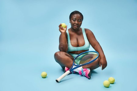 Photo for Young plus size dark skinned sportswoman holding tennis racket and sitting around balls on blue - Royalty Free Image