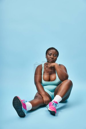 Photo for Relaxed plus size african american sportswoman sitting in blue sportswear on matching backdrop - Royalty Free Image
