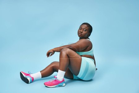 Photo for Relaxed plus size african american sportswoman sitting in blue sportswear on matching background - Royalty Free Image