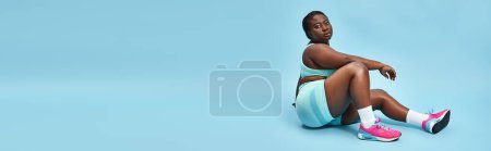 Photo for Relaxed plus size african american woman sitting in blue sportswear on matching backdrop, banner - Royalty Free Image