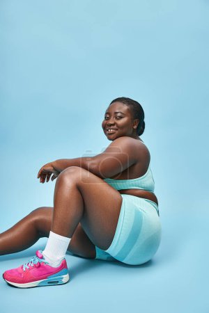Photo for Cheerful plus size african american woman sitting in blue sportswear on matching backdrop, sport - Royalty Free Image