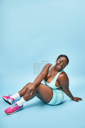 Photo for Happy plus size african american woman sitting in blue sportswear on matching backdrop, sport - Royalty Free Image