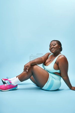 Photo for Smiling plus size african american woman sitting in blue sportswear on matching backdrop, sport - Royalty Free Image