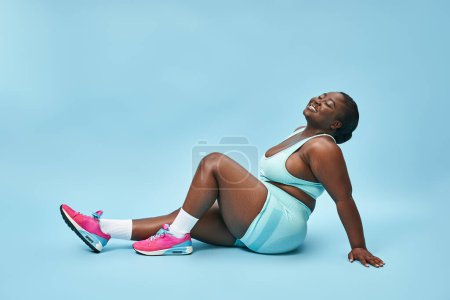 Photo for Relaxed plus size african american woman sitting in blue sportswear on matching backdrop, smile - Royalty Free Image