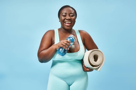 Lächelnde plus size afrikanisch-amerikanische Frau steht mit Fitnessmatte und Wasserflasche auf blau