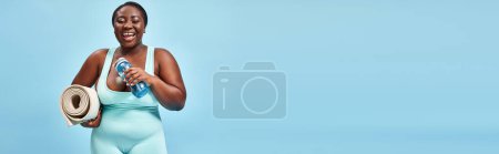 cheerful plus size african american woman standing with fitness mat and water bottle on blue, banner