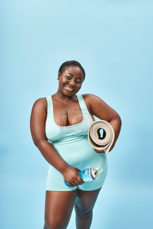 freudige plus size afrikanisch-amerikanische Frau steht mit Fitnessmatte und Wasserflasche auf blau