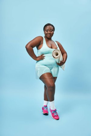 Smiling plus size african american woman standing with fitness mat on blue backdrop, sport